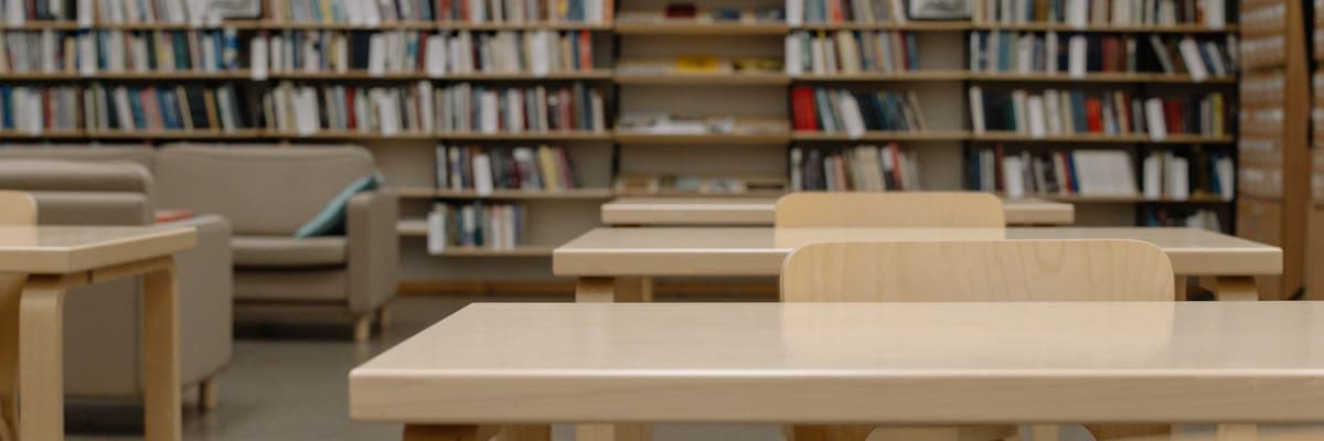 Wooden study tables and chairs in a library, with sofas and long shelves of books along the back wall.