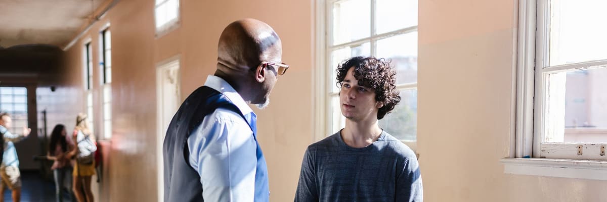 A teacher in a vest talking with a student who holds a music book, in a sunlit school hallway, the setting where a school psychologist works.
