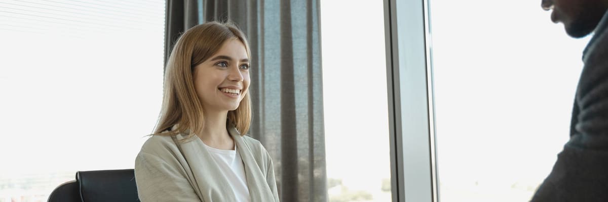 A smiling young woman in a light jacket shaking hands with a man in a dark sweater beside a sunlit office window, the kind of meeting an APPIC internship application leads to.
