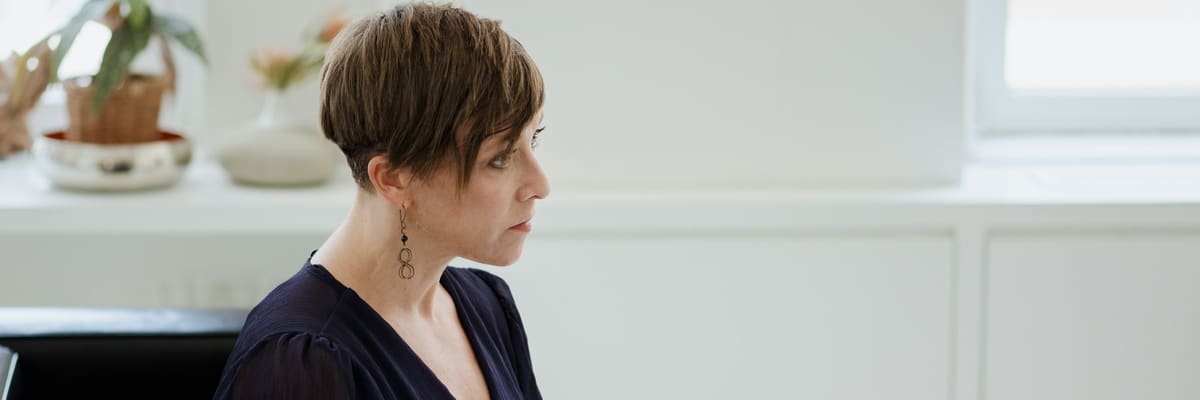 A woman with short brown hair in a navy blouse sits in a bright room beside a window, holding a pen and a clipboard and looking to one side as she works out how to become a licensed psychologist.