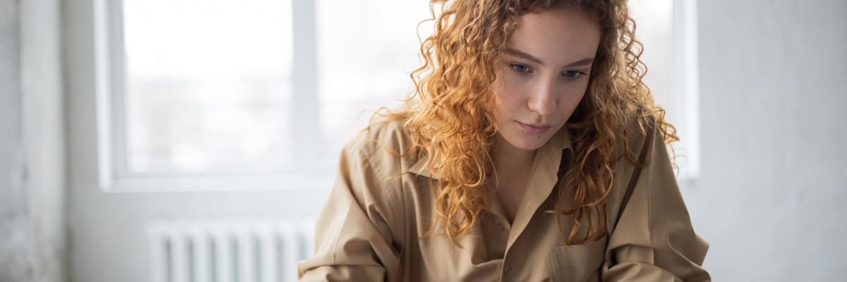 A woman with curly red hair writing in a notebook at a dark table, with an open textbook in front of her and a window behind, studying how to become a psychologist.