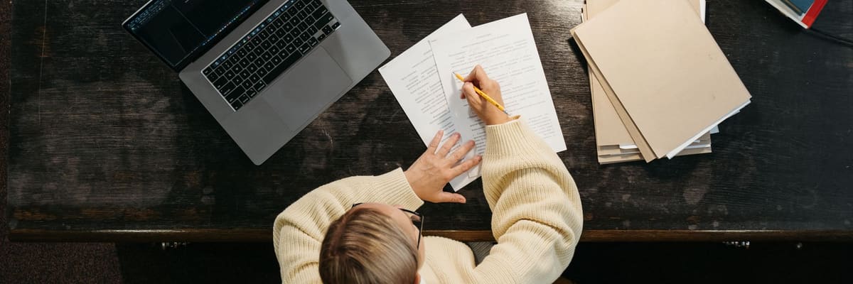 PsyD requirements start at a desk: seen from above, a person in a cream sweater marking up papers beside an open laptop and stacked folders.