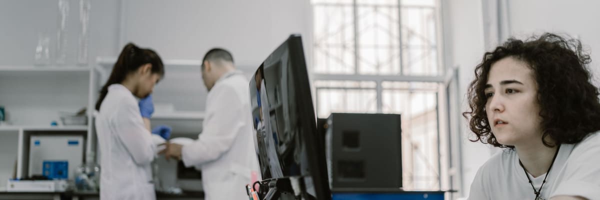 A student working at a computer in a research laboratory, with two colleagues in lab coats at a bench behind her, the research side of the PsyD vs PhD comparison.