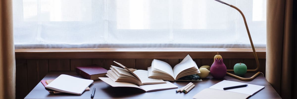 A study desk in front of a sheer-curtained window, with open books, notebooks, pencils and a gold desk lamp.