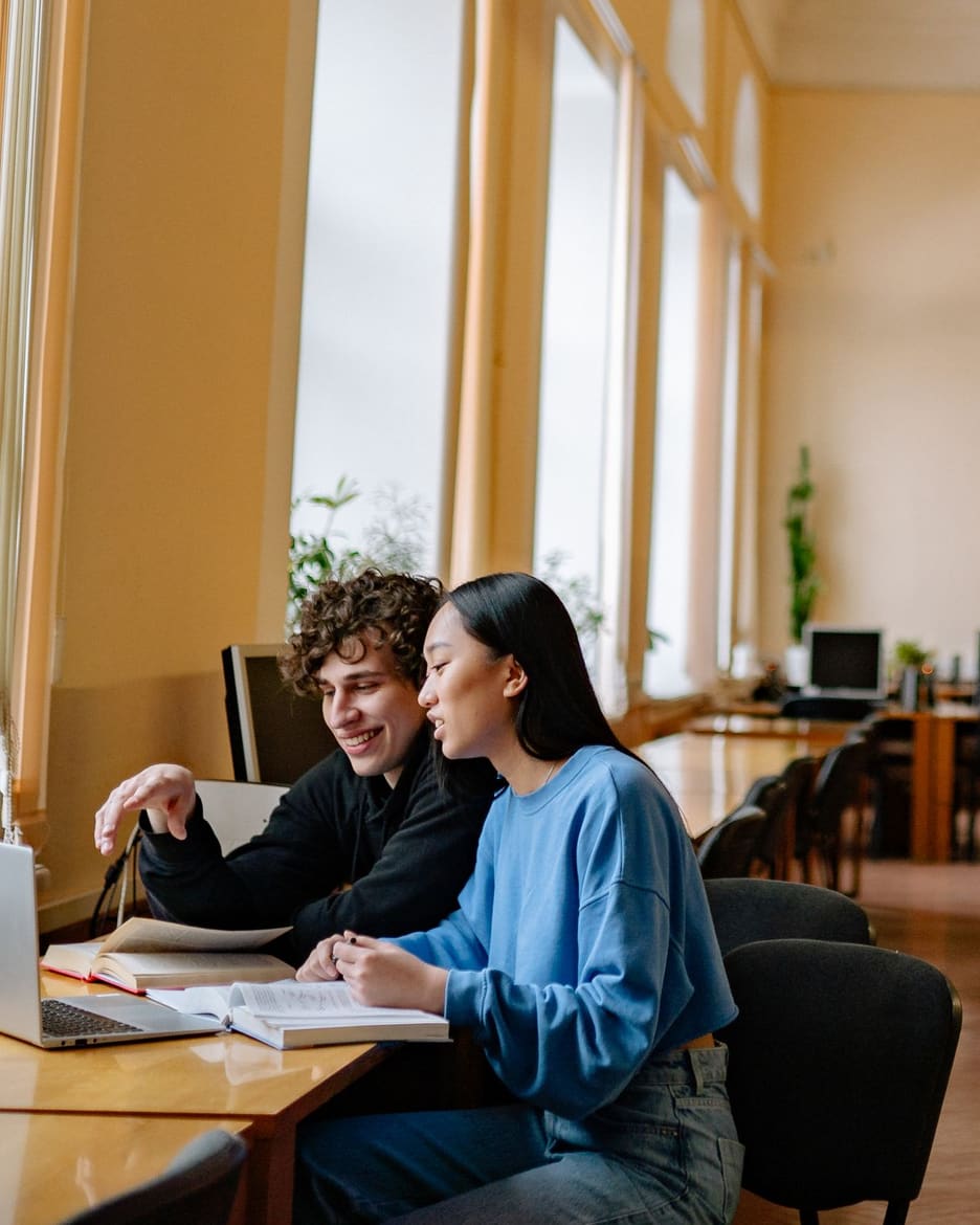 Two graduate students at a library table beside tall arched windows, one pointing at an open textbook and the other writing notes, the study that goes into choosing among PsyD programs.