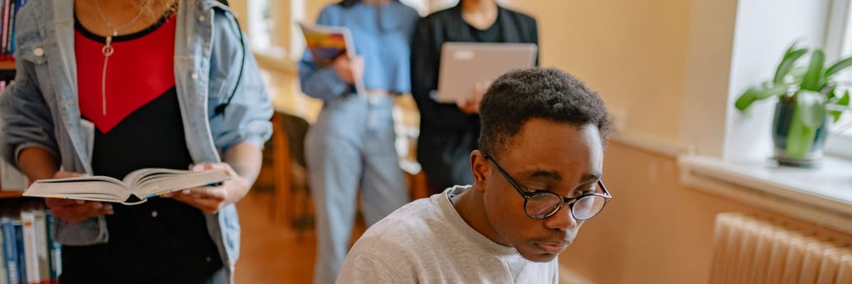 A graduate student working at a library desk while classmates carry books behind him, a study scene for the best PsyD programs.