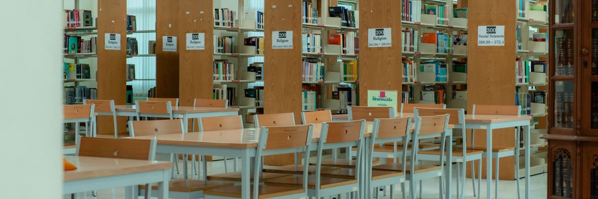Empty study tables and chairs in a bright library between tall shelves of books, a quiet place to study for counseling psychology doctoral programs.