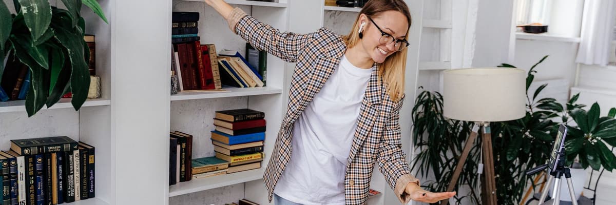 A woman in a plaid blazer gesturing toward a phone on a tripod in a home study with full bookshelves, for online PsyD programs.