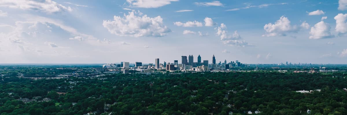 The Atlanta, Georgia skyline rising above a canopy of green trees under a bright blue sky with scattered clouds. The photograph opens the ranking of PsyD programs in Georgia.