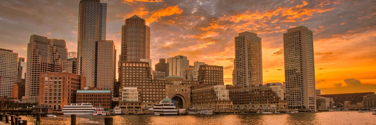 Downtown Boston, Massachusetts, towers and the Rowes Wharf arch glowing across the harbor under an orange sunset sky. The photograph opens the ranking of PsyD programs in Massachusetts.
