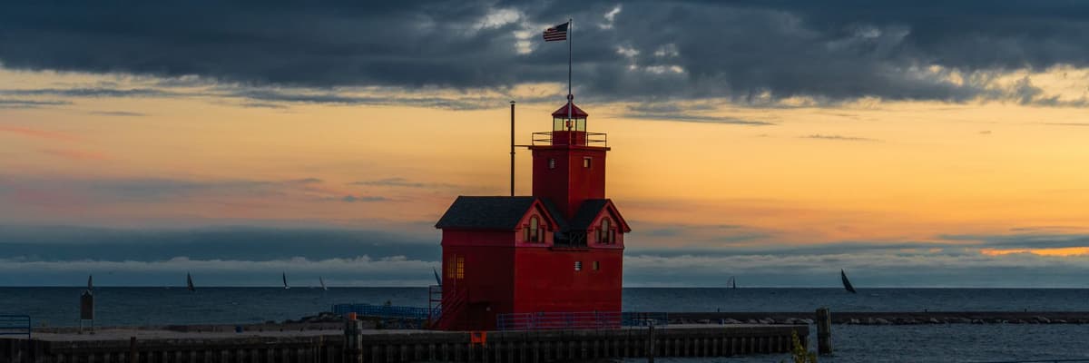 The red Holland Harbor Lighthouse on its pier at dusk under dark clouds, with sailboats on the horizon of Lake Michigan, in Holland, Michigan. The photograph opens the ranking of PsyD programs in Michigan.