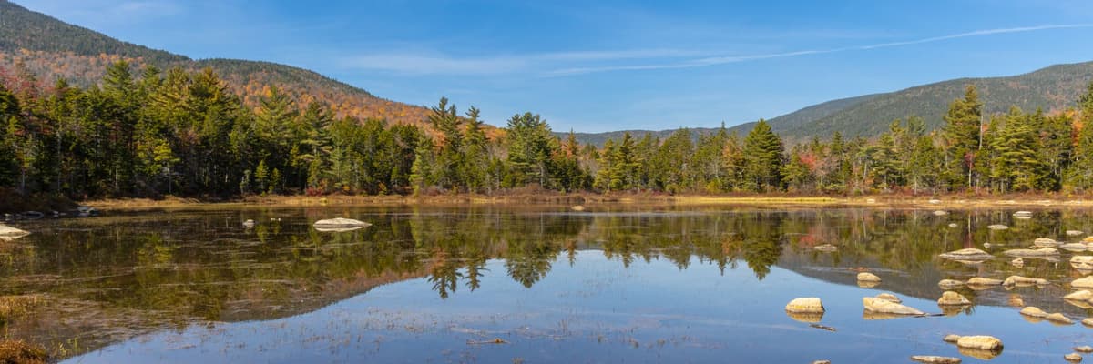 A still pond reflecting evergreens and autumn color below forested mountains in Franconia, New Hampshire. The photograph opens the ranking of PsyD programs in New Hampshire.