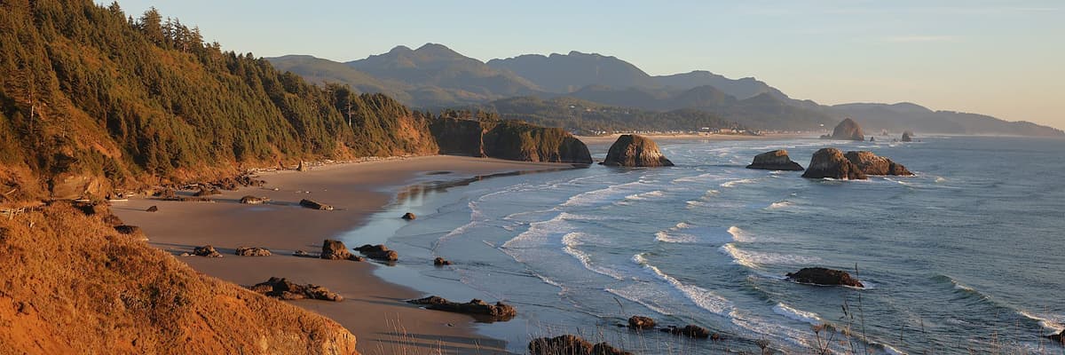 Waves rolling onto a sandy beach below forested headlands and sea stacks near Cannon Beach on the Oregon coast. The photograph opens the ranking of PsyD programs in Oregon.