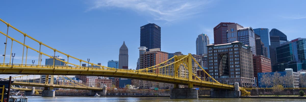 A yellow suspension bridge over a river with the downtown Pittsburgh, Pennsylvania skyline behind it under a blue sky. The photograph opens the ranking of PsyD programs in Pennsylvania.