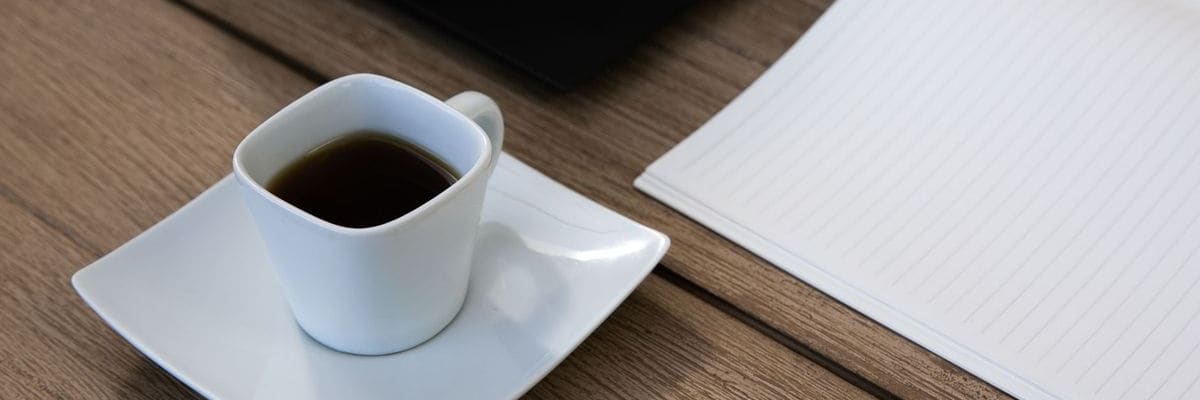 A small white cup of black coffee on a saucer beside an open lined notebook on a wooden desk.
