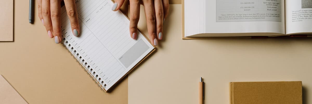 The psychology internship match takes planning: seen from above, a pair of hands resting on a spiral planner beside an open book, a pencil and a notebook on a tan desk.