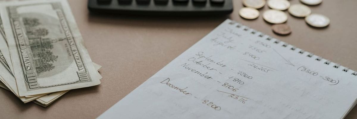 PsyD tuition adds up: hundred-dollar bills fanned out beside a spiral notebook of handwritten monthly figures, with a calculator and coins at the top of the desk.