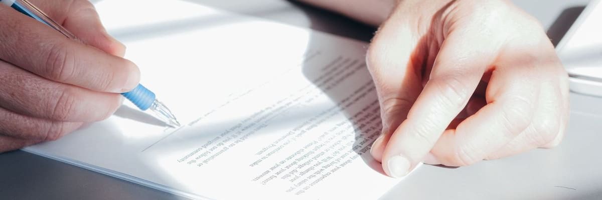 A hand signing a printed document with a blue pen while the other hand rests on the page.